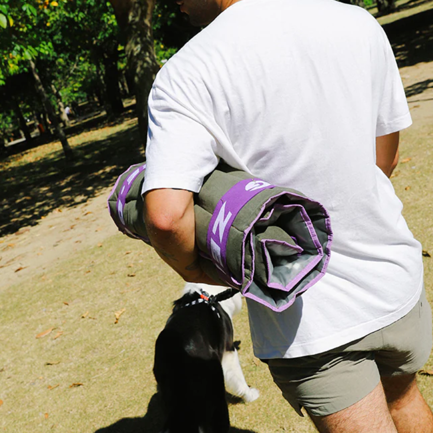 Person holding a rolled-up air mat with a logo, walking a dog on a path.
